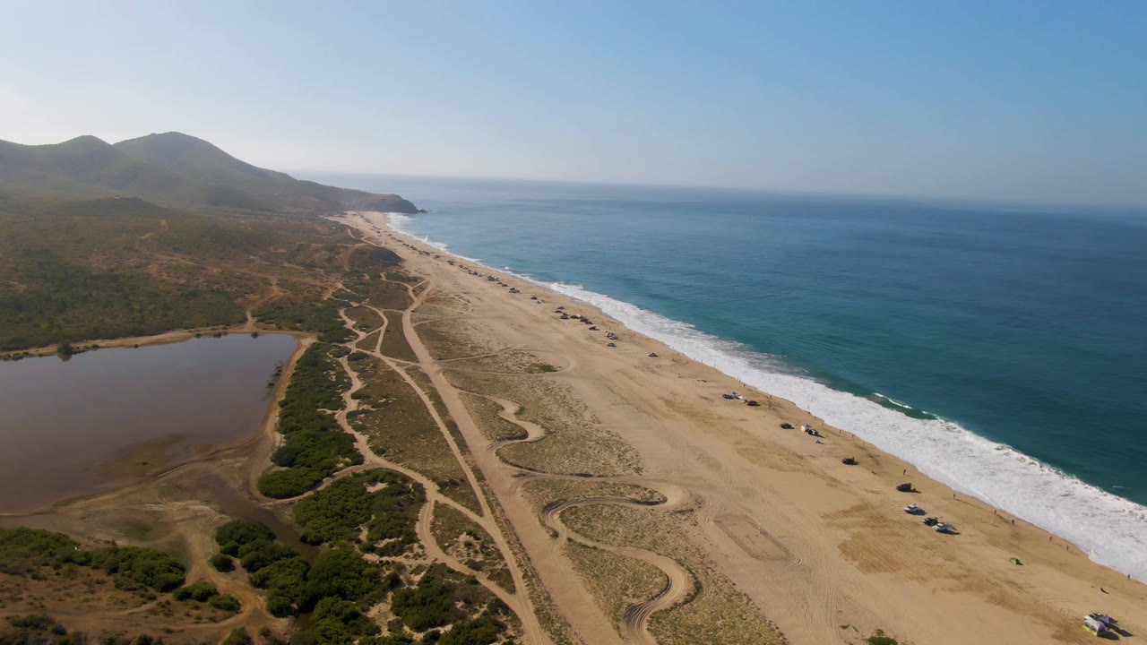 Drone view of sandy beach, lagoon, ocean surf, and distant mountains.