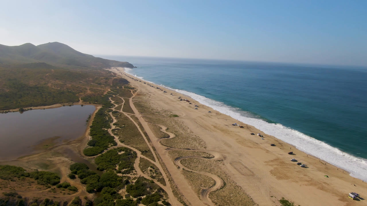 At 0:01 the Migrino aerial view holds the coastline diagonally across frame, with the lagoon lower left, surf breaking along the beach, distant hills, and no visible people.