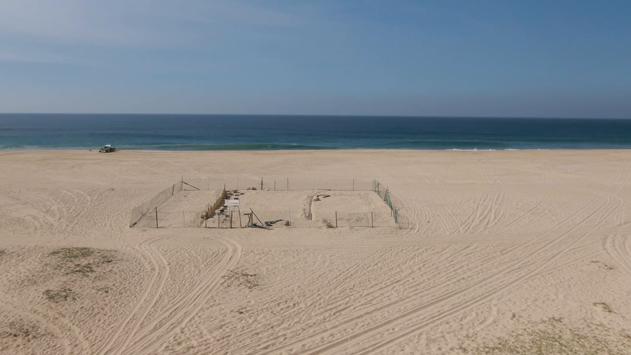 Wide sandy beach with fenced area, ocean horizon, and small vehicle near the surf.