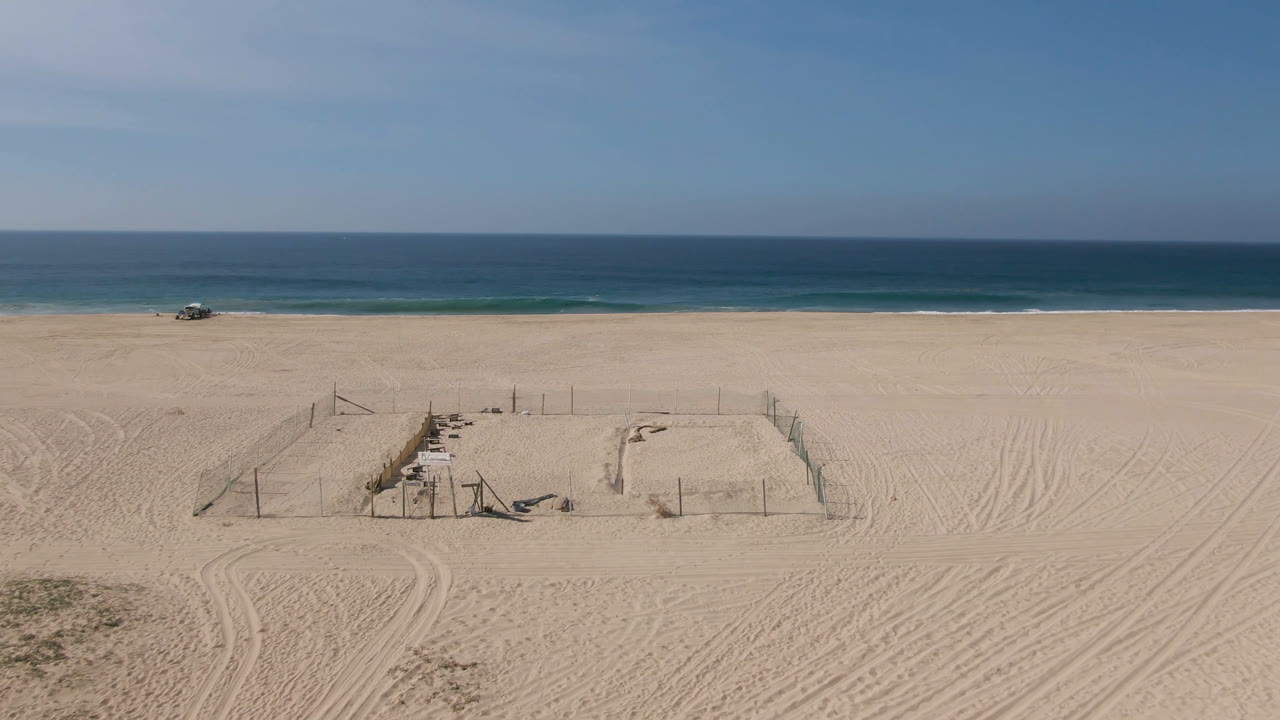 Aerial-style frame over Migrino beach showing tire-marked sand, a fenced rectangle near the lower center, a small vehicle close to the shoreline, blue water, and no visible people.