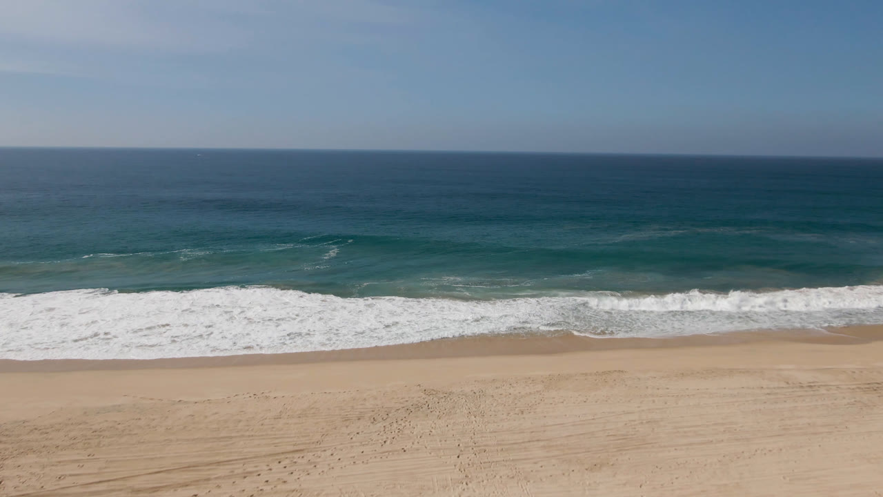 Opening aerial frame from Migrino, Baja California Sur, Mexico, showing white surf rolling toward tan sand, teal ocean beyond, a flat horizon, and no visible people.