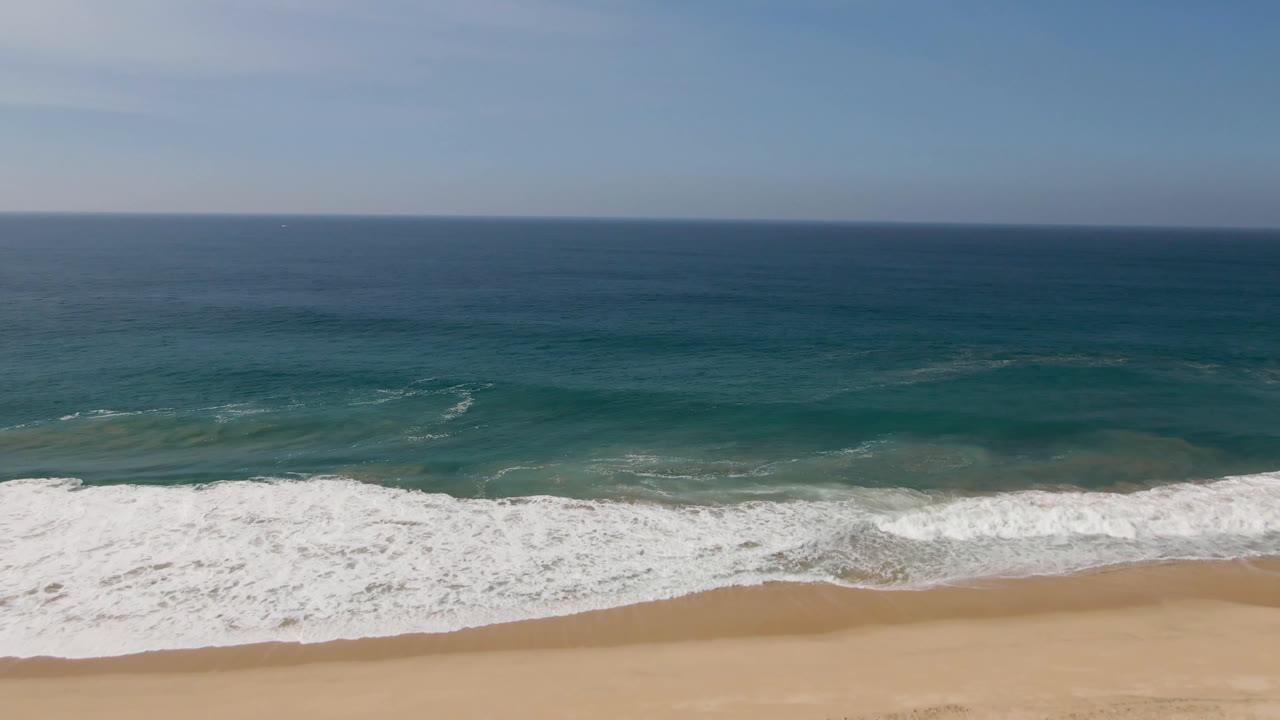 Aerial coastal frame in Migrino, Baja California Sur, Mexico, with a wider band of white water over the sand, blue-green ocean in the middle distance, and no visible people.