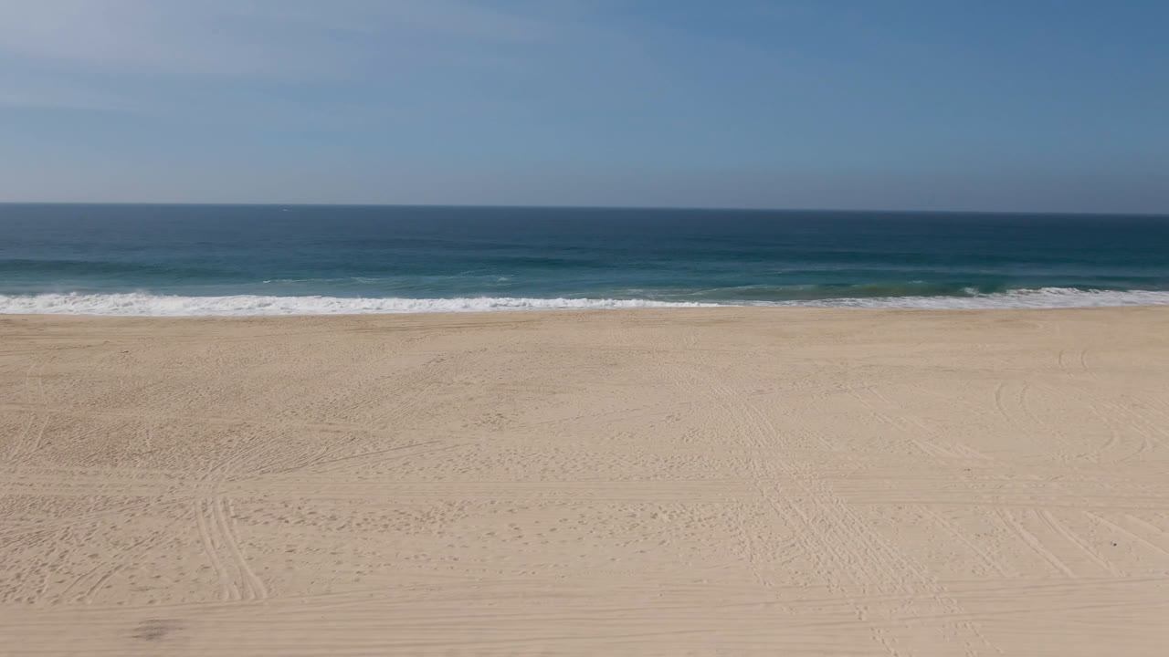 Elevated view across sunlit Migrino sand toward turquoise ocean bands and low white waves, with no visible people in the frame.