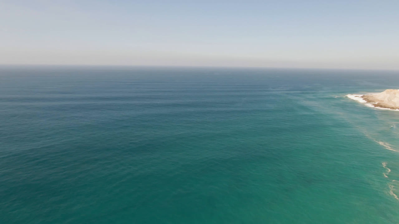 Aerial view over turquoise ocean near Migrino, Baja California Sur, with pale sand and low coastal cliff entering from the upper right and no visible people.