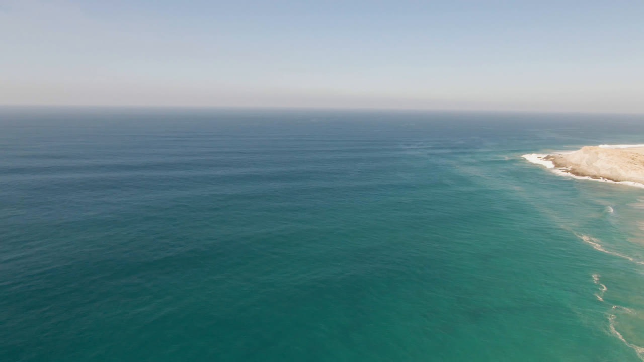 Wide aerial frame near Migrino, Baja California Sur, showing layered blue water, faint surf streaks, and a sandy point along the right side with no visible people.