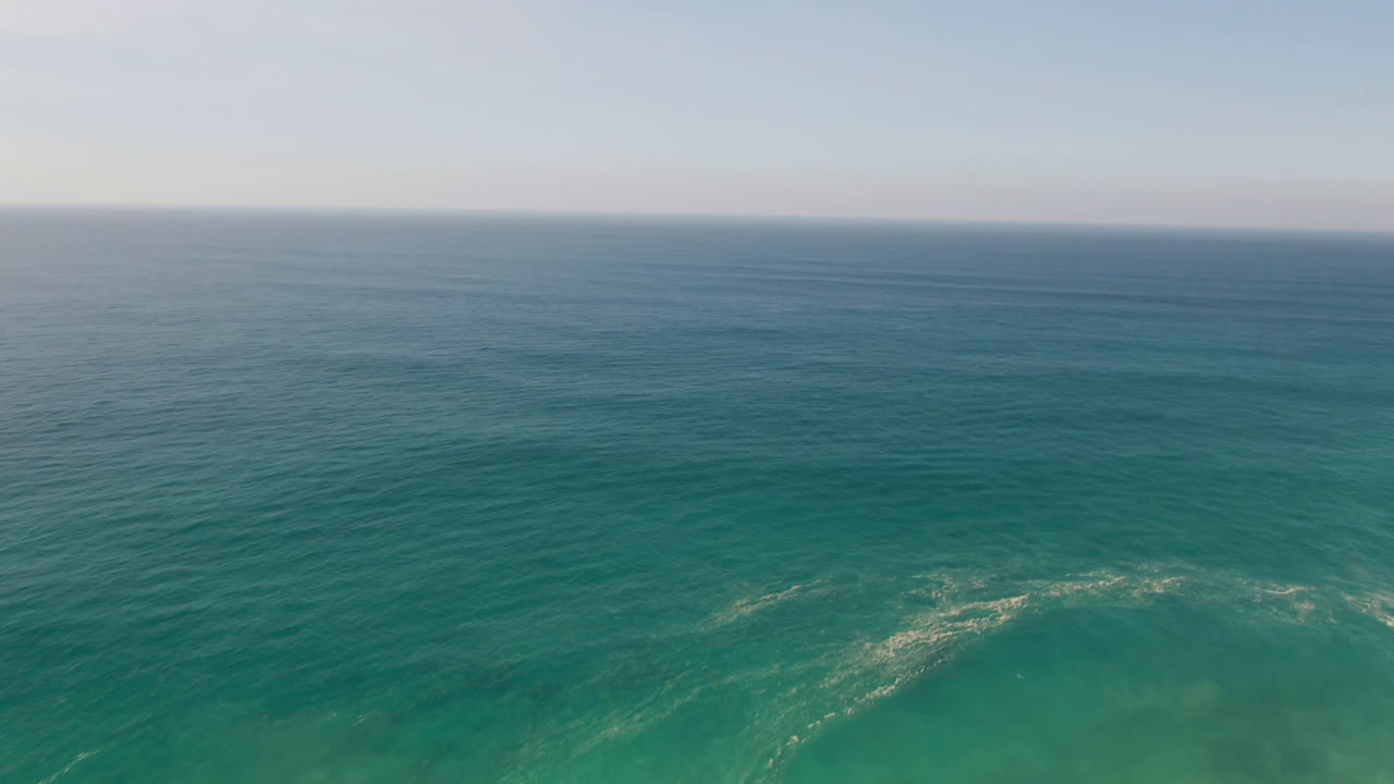 Aerial view over teal ocean water in Migrino, Baja California Sur, Mexico, with a level horizon, pale blue sky, faint surf texture near the bottom, and no visible people.