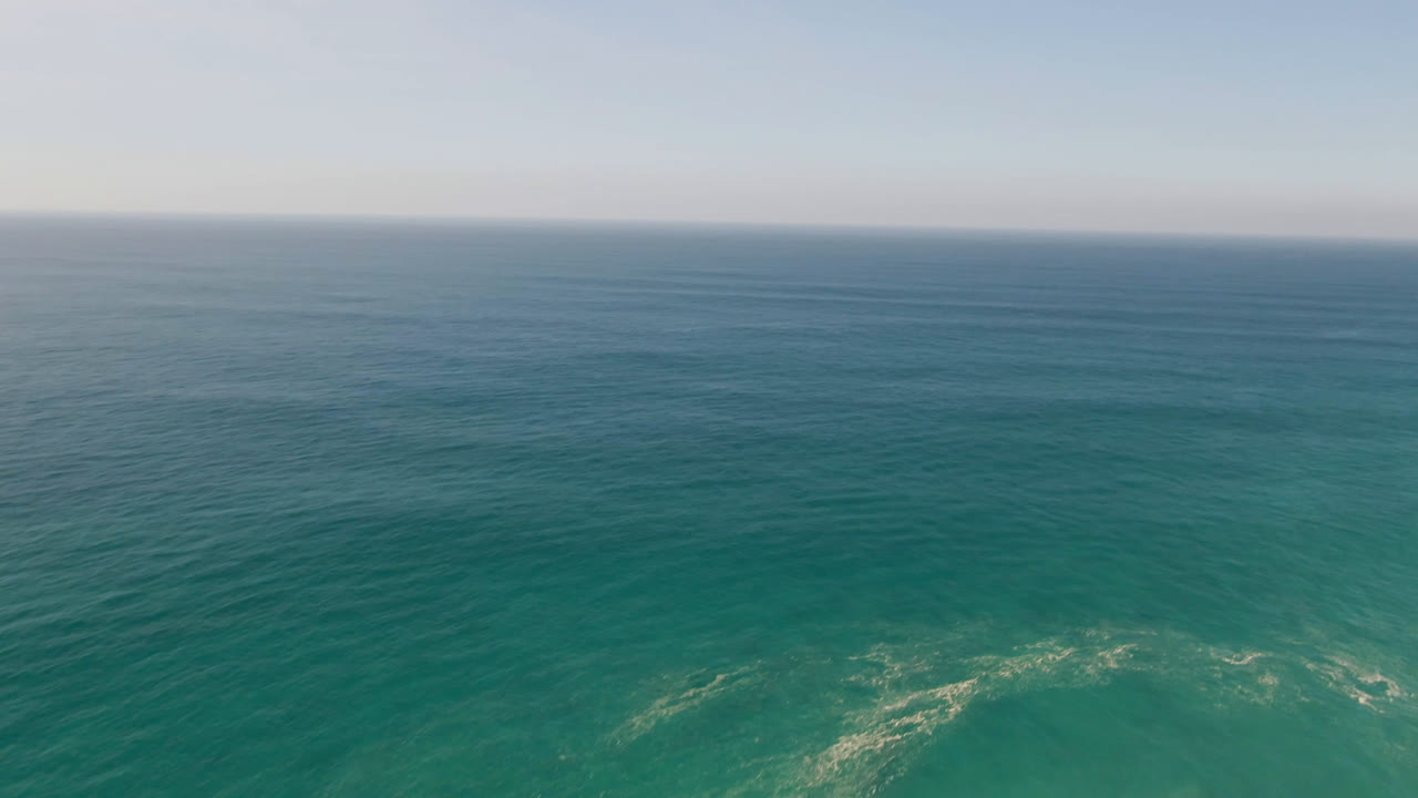 Aerial frame over open teal water in Migrino, Baja California Sur, Mexico, showing subtle wave bands, a pale sky, a distant horizon, and no visible people.