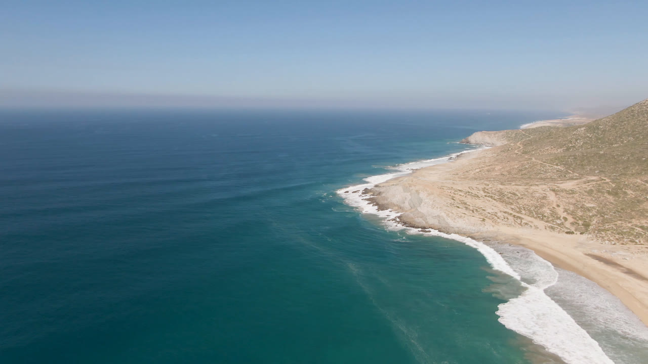 Aerial view over Migrino, Baja California Sur showing blue ocean, a curving sandy beach, white surf, dry cliff terrain, and no visible people at the start of the clip.