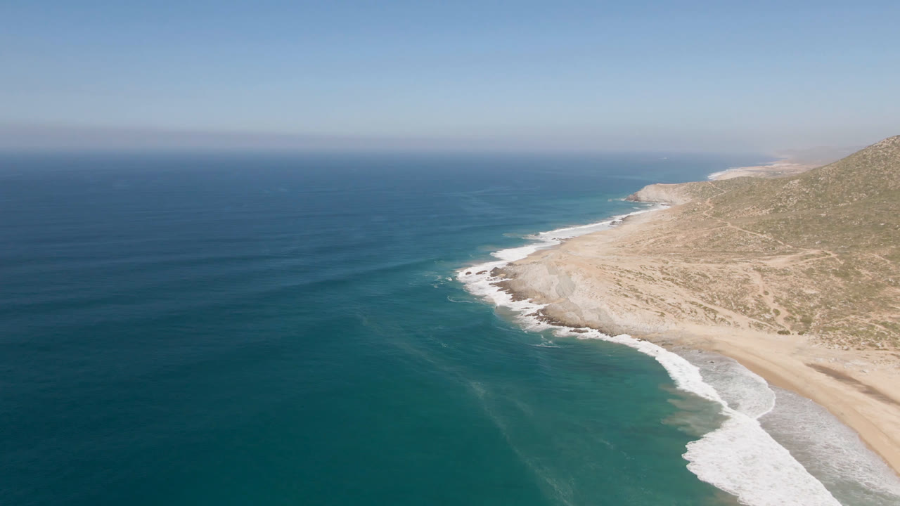Wide aerial frame over Migrino, Baja California Sur with the shoreline curving below a dry hillside, darker ocean water offshore, and no visible people.