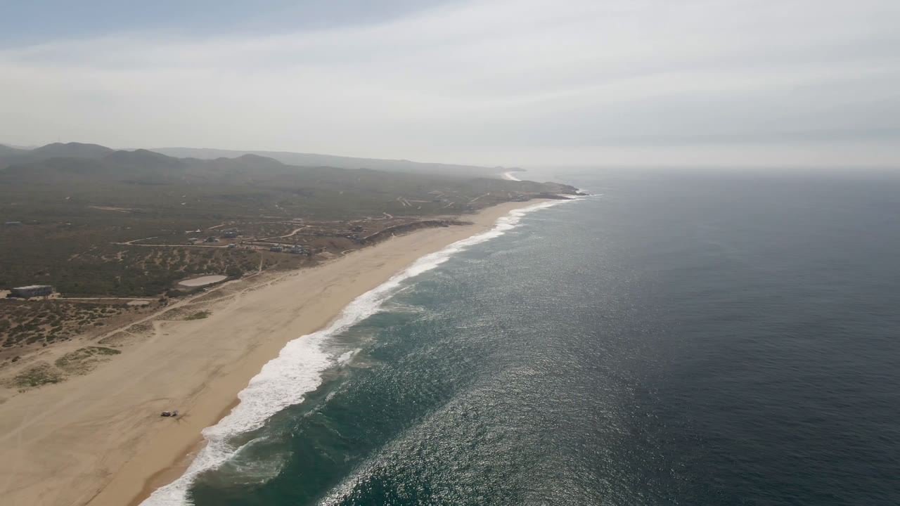Opening aerial frame over Migrino, Baja California Sur, with tan beach at left, dark ocean at right, white surf along the diagonal shore, low hills inland, and no visible people.