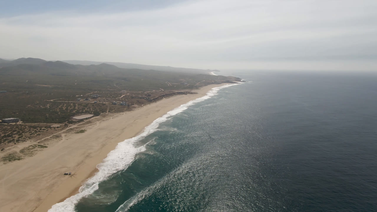 High coastal aerial with beach, foamy surf, ocean surface, inland terrain, and gray clouds.