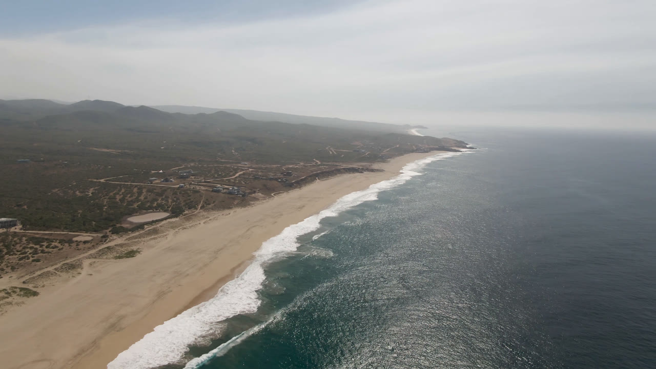 Opening aerial frame over Migrino, Baja California Sur, showing a wide sandy beach, foamy surf, dark blue ocean, inland scrub roads, distant hills, and no visible people.