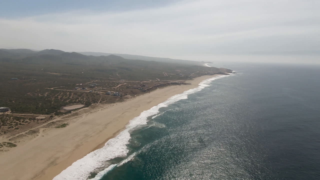 Aerial view of Migrino beach one second in, with the shoreline angled through the frame, waves breaking in bands, inland tracks and scrub visible, and no visible people.