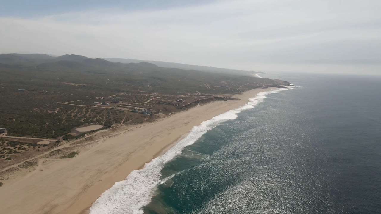 Aerial frame over Migrino, Baja California Sur showing a long sandy beach, white surf on the shoreline, blue-green ocean to the right, inland scrub and hills, and no visible people.