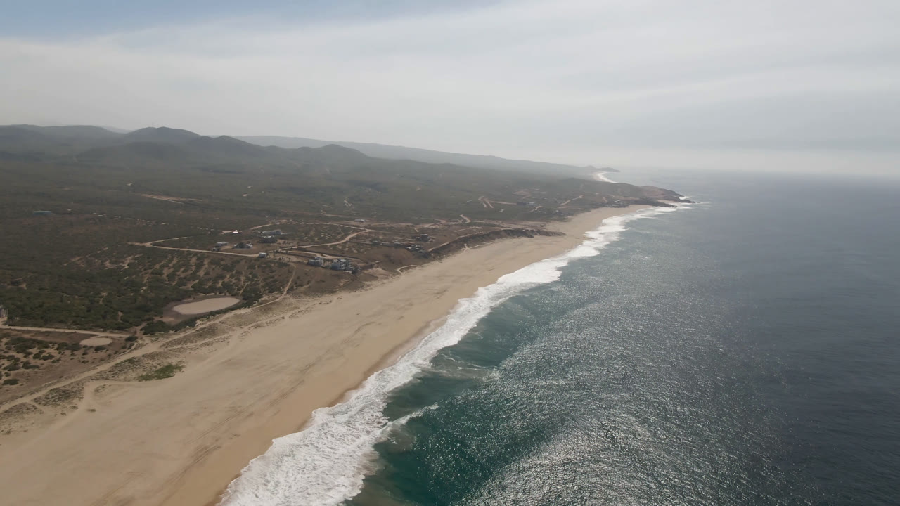 Aerial view in Migrino, Baja California Sur with the beach running diagonally through frame, repeating surf bands, inland dirt roads and scrubland, distant hills, and no visible people.