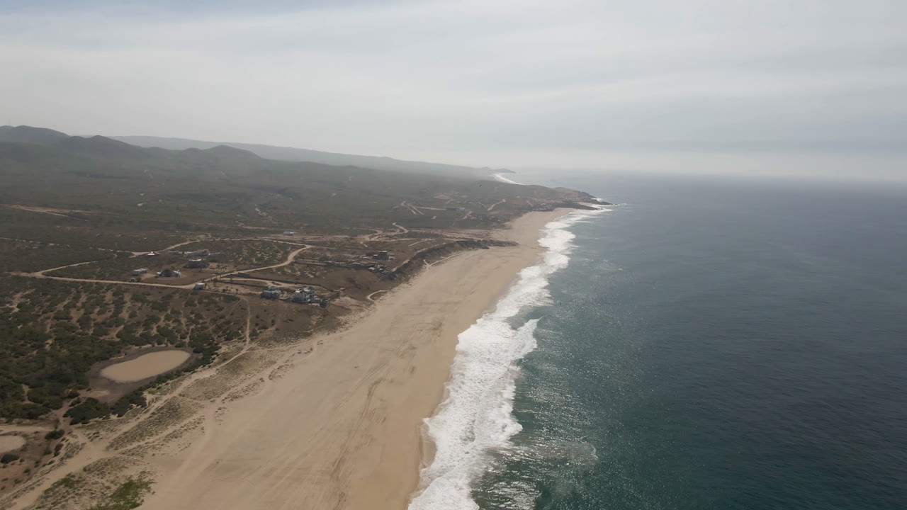 Aerial view over Migrino, Baja California Sur, with a wide sand beach meeting rolling surf, dry inland roads and low hills to the left, ocean to the right, and no visible people.