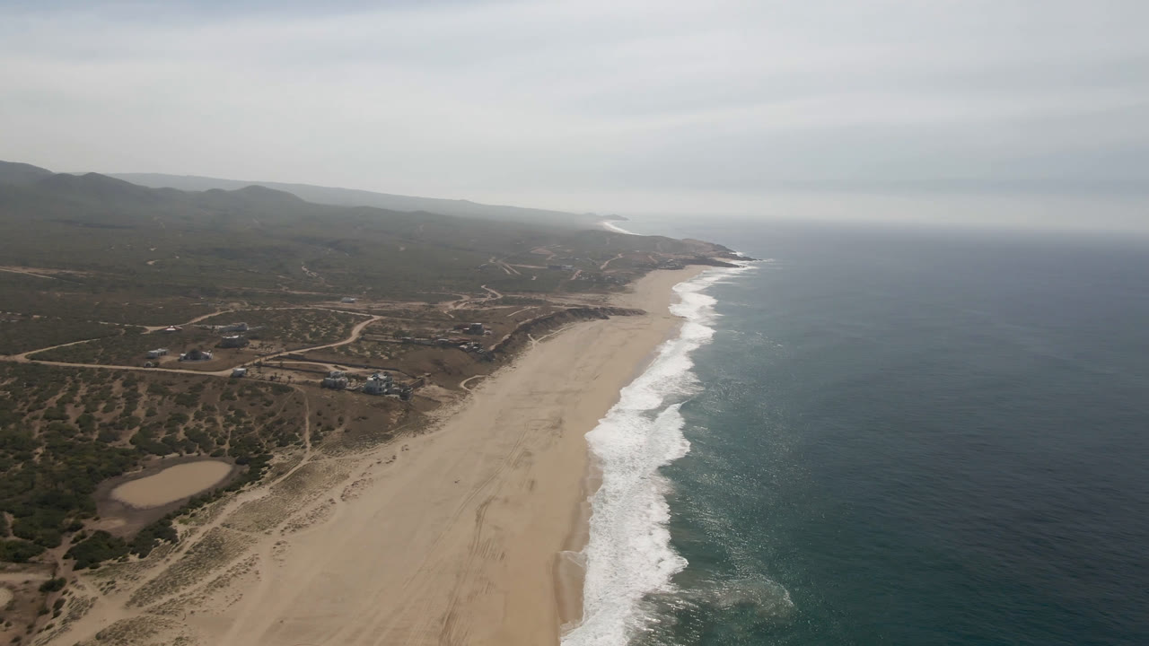 Drone view of long beach, ocean surf, inland tracks, and hills under clouds.
