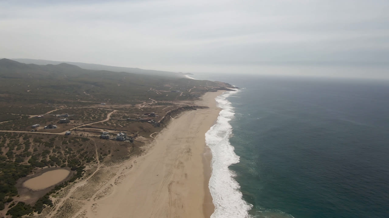 High aerial view over Migrino, Baja California Sur, with a sandy beach below, white surf along the shore, ocean to the right, inland roads and hills to the left, and no visible people.