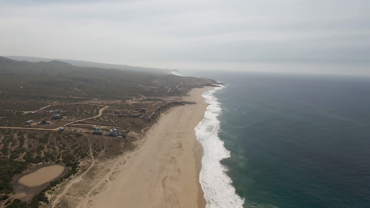 Aerial frame over Migrino coastline showing the beach stretching into the distance, waves breaking along the right edge, winding dirt roads inland, low hills on the horizon, and no visible people.