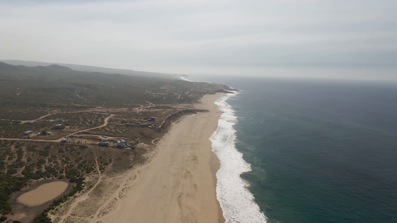 High coastal aerial with ocean, foamy waves, broad sand, and inland hills.