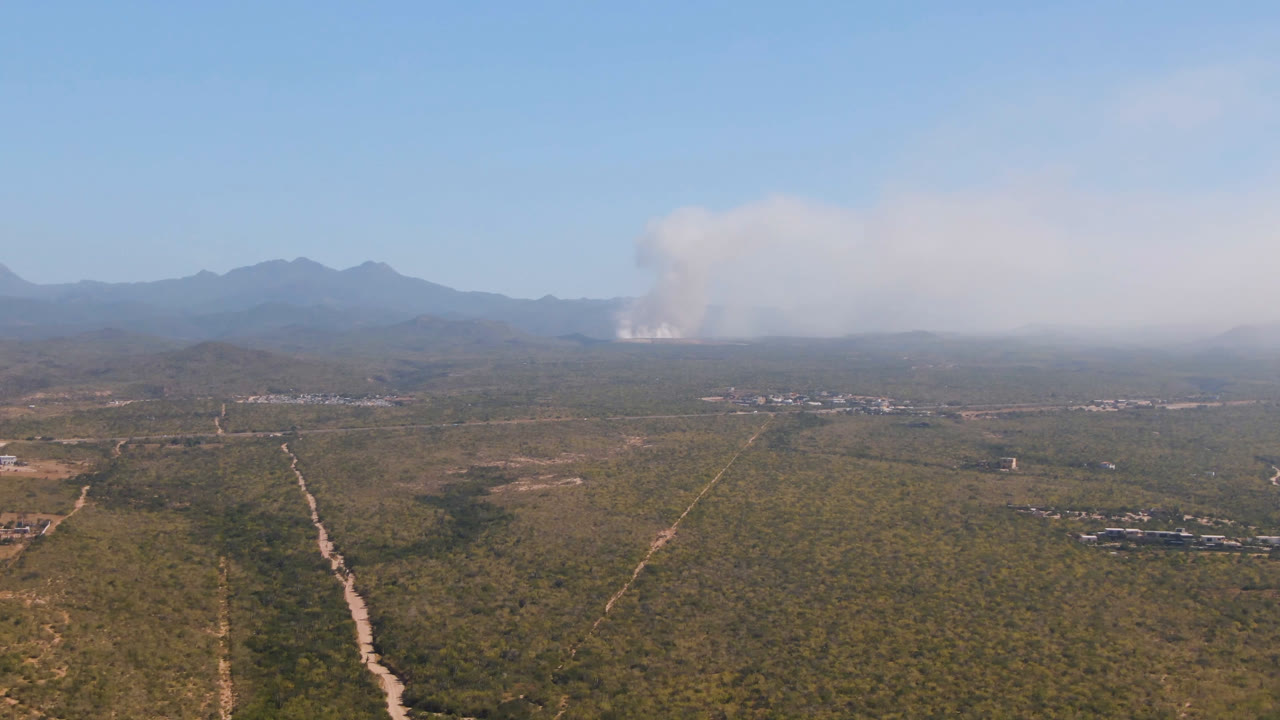 Aerial scrubland with dirt roads and distant smoke plume near mountains