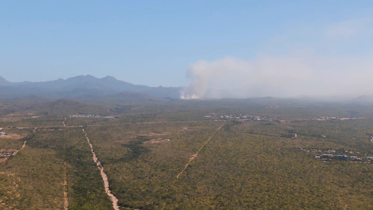 Wide aerial frame over Migrino, Baja California Sur, with green-brown terrain, a central dirt road, low hills, distant mountains, and smoke thickening across the horizon with no visible people.