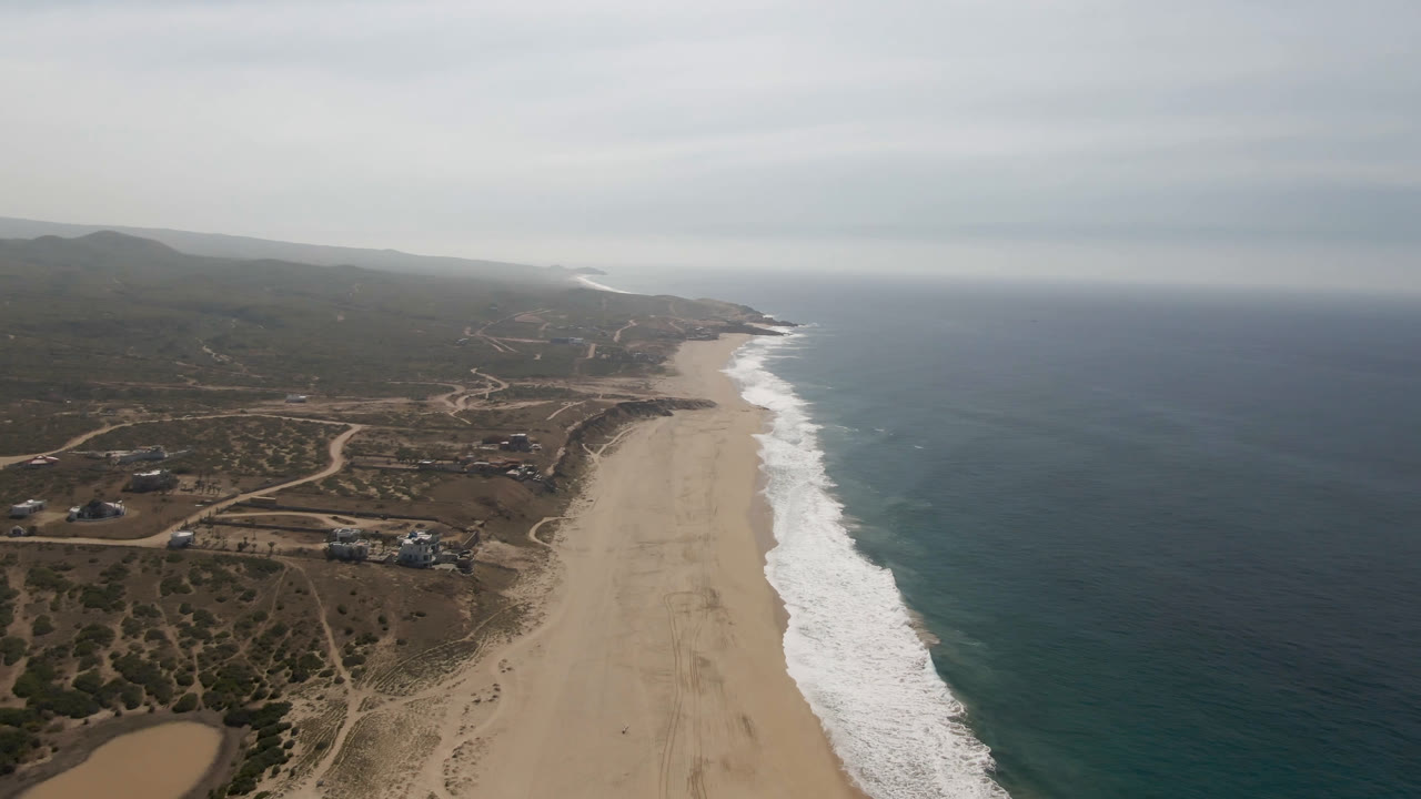 Opening aerial frame over Migrino, Baja California Sur, with a wide sandy beach below, white surf on the right, dirt tracks and scattered buildings inland, and no clearly recognizable people.