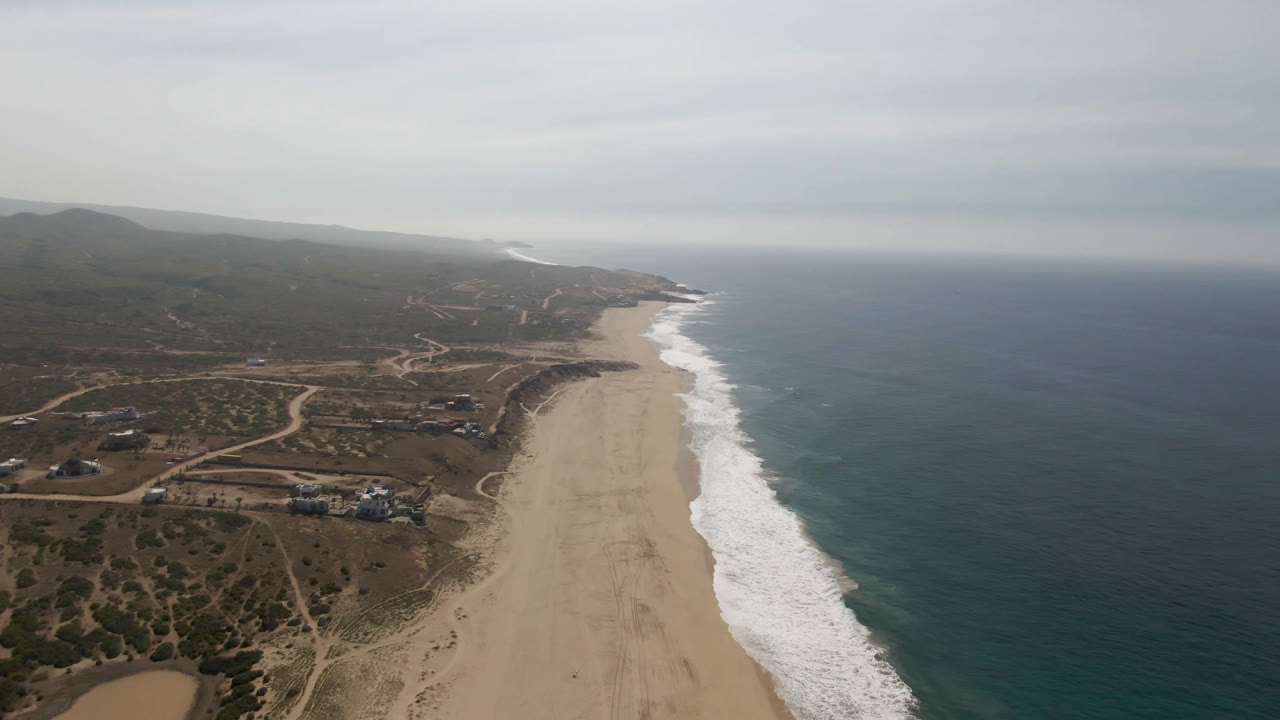 Aerial view along the Migrino coastline with the beach stretching toward the distance, ocean surf forming a white edge, inland dirt roads visible, and no clearly recognizable people.
