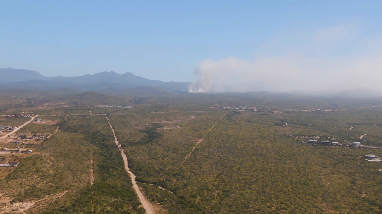 Aerial view of desert roads, scattered buildings, mountains, and distant smoke.