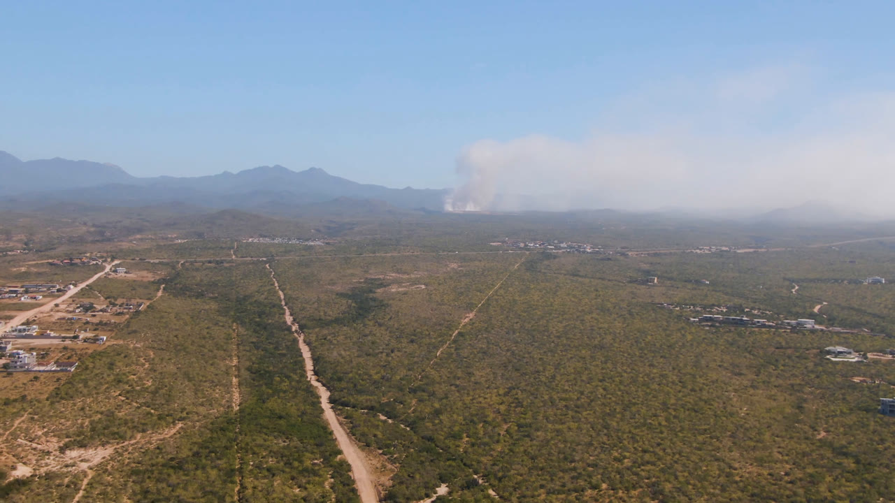 Aerial view in Migrino, Baja California Sur, with a pale dirt road running from the foreground into scrubland, small structures along the left edge, distant mountains, smoke, and no visible people.