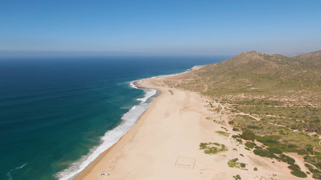 Opening aerial frame over Migrino, Baja California Sur, showing blue ocean, pale beach, low coastal scrub, rocky hills, and no visible people.