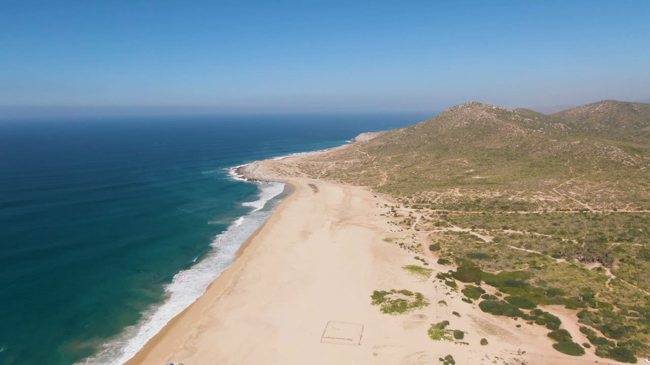 Aerial frame from Migrino, Baja California Sur, with white wave bands along the left edge, broad pale sand in the center, green scrub patches, hills, and no visible people.