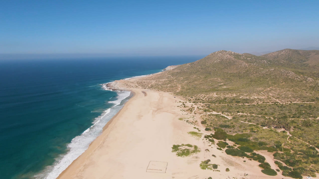 High coastal aerial with surf, empty sand, scrubland, and a hill ridge.