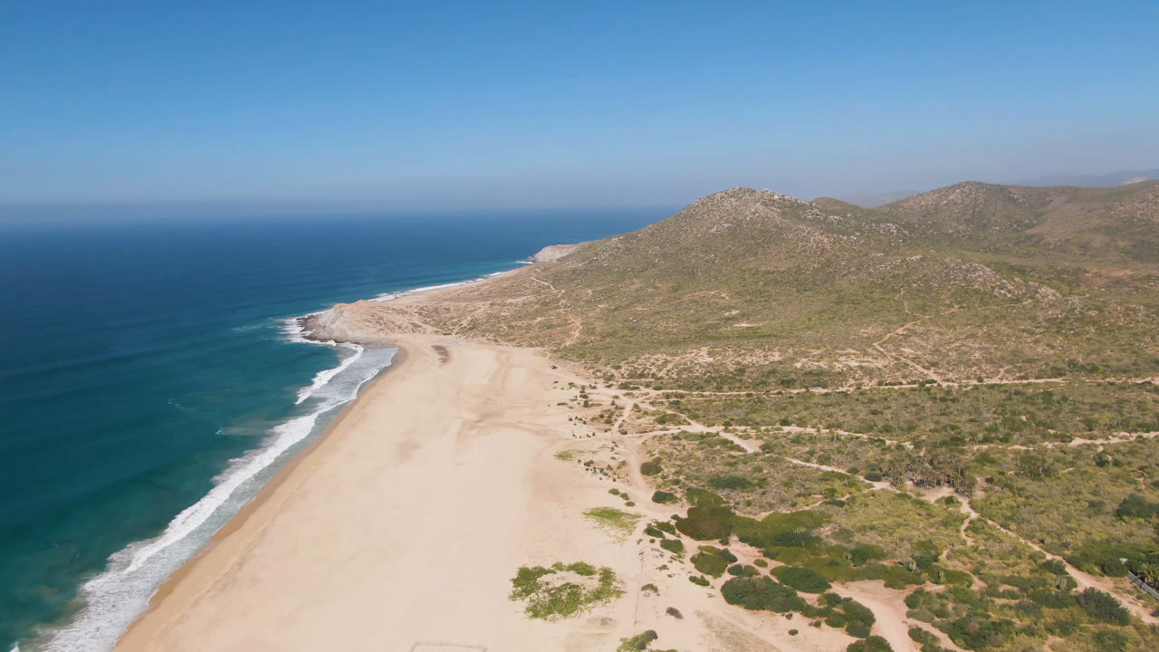Aerial frame over Migrino, Baja California Sur, with blue ocean on the left, a broad pale beach below, scrubland and rocky hills beyond, and no visible people.