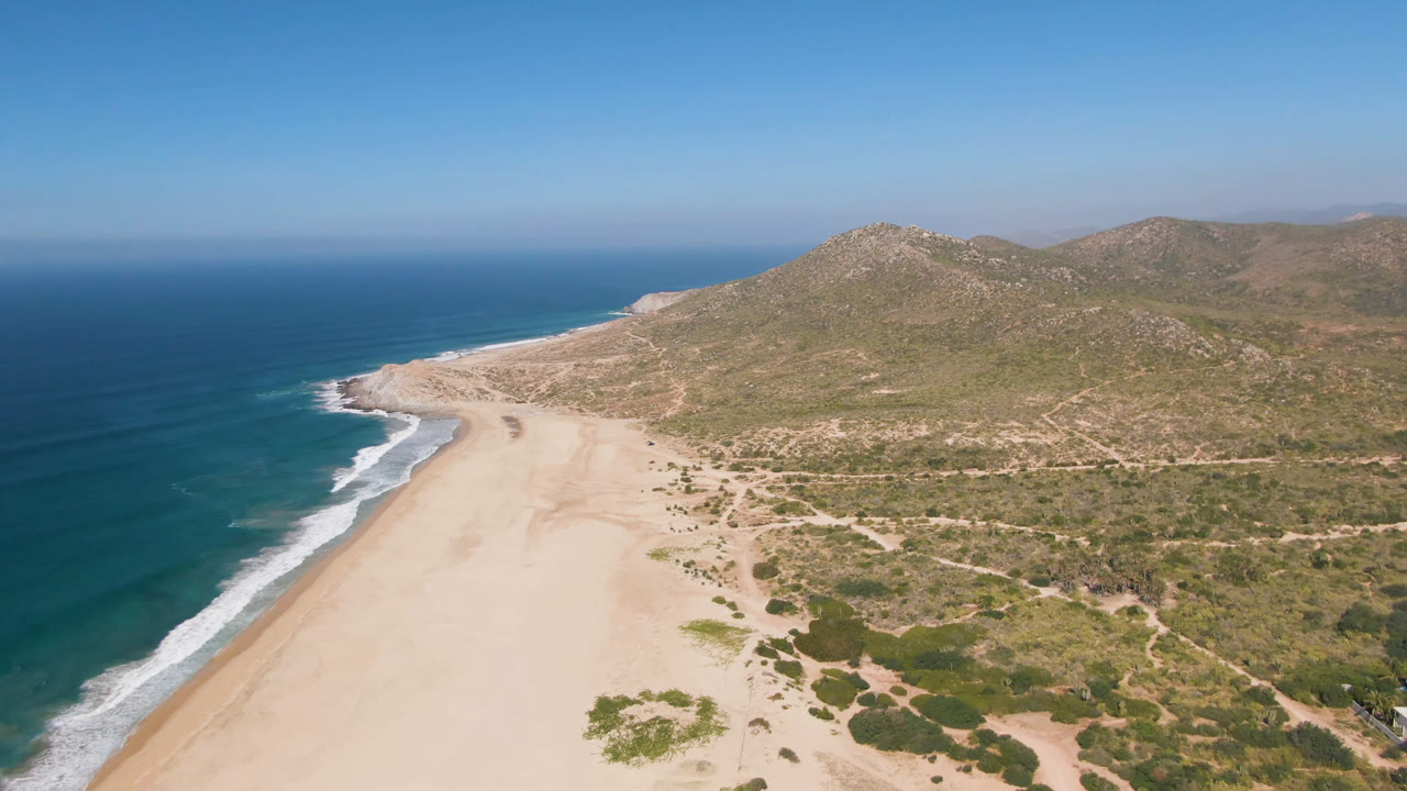 Aerial view in Migrino, Baja California Sur, showing the shoreline curving beside deep blue water, with dry scrub and low hills filling the inland side and no visible people.
