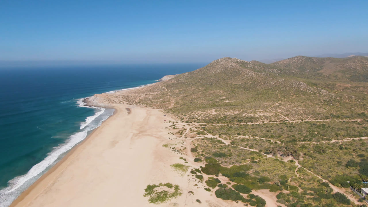 Wide coastal aerial with beach, waves, scrub-covered land, and mountains