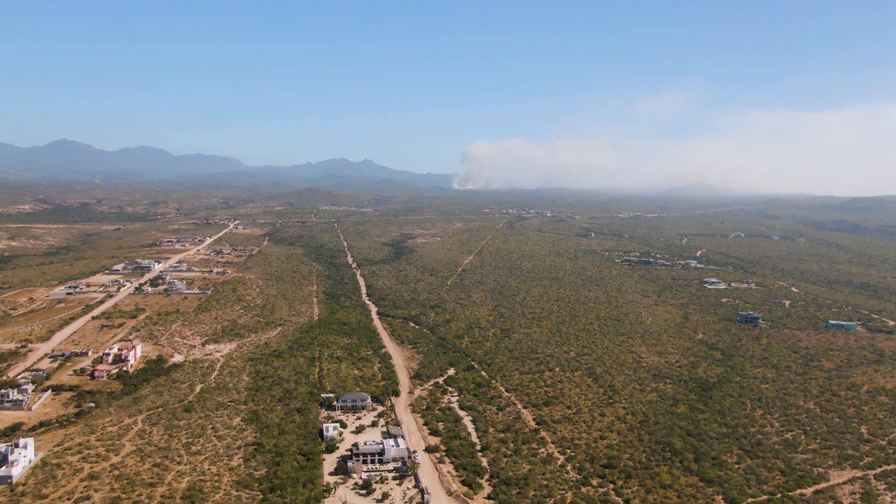 Aerial view of desert roads, scattered buildings, distant mountains, and smoke near the horizon.