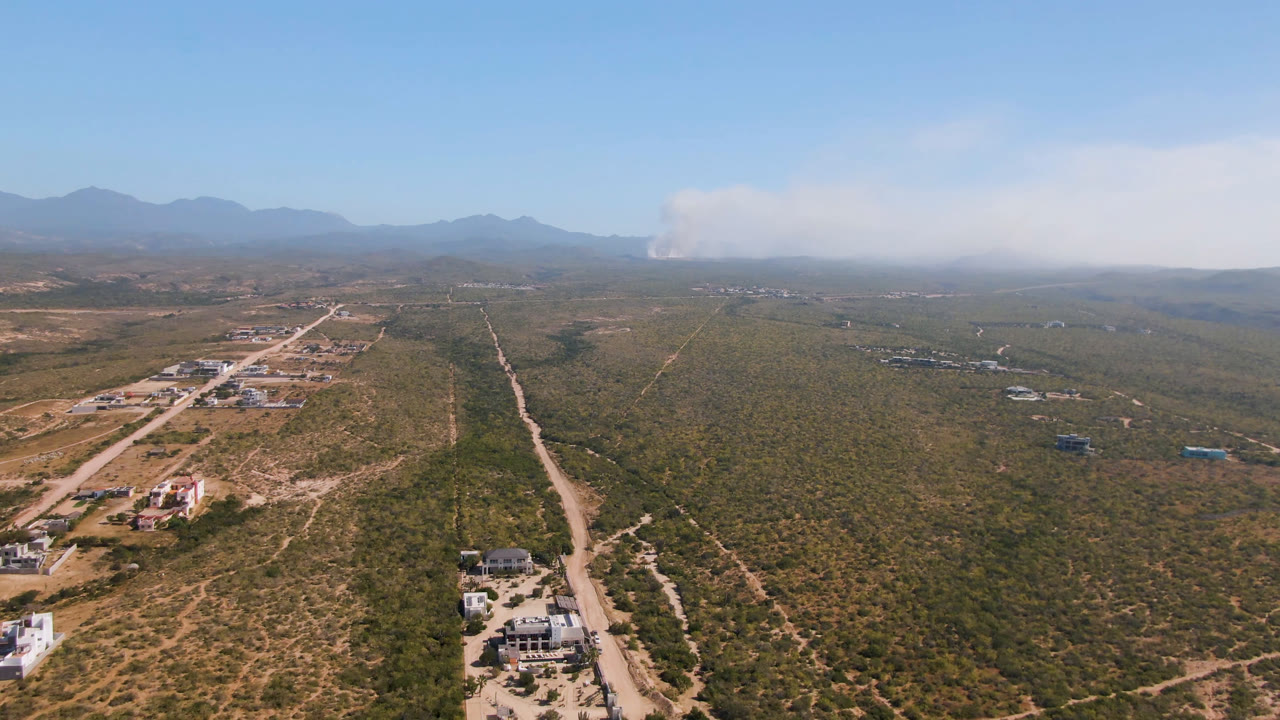 Early aerial frame in Migrino, Baja California Sur with a long dirt road through scrubland, low buildings below, mountains in the distance, and no visible people.