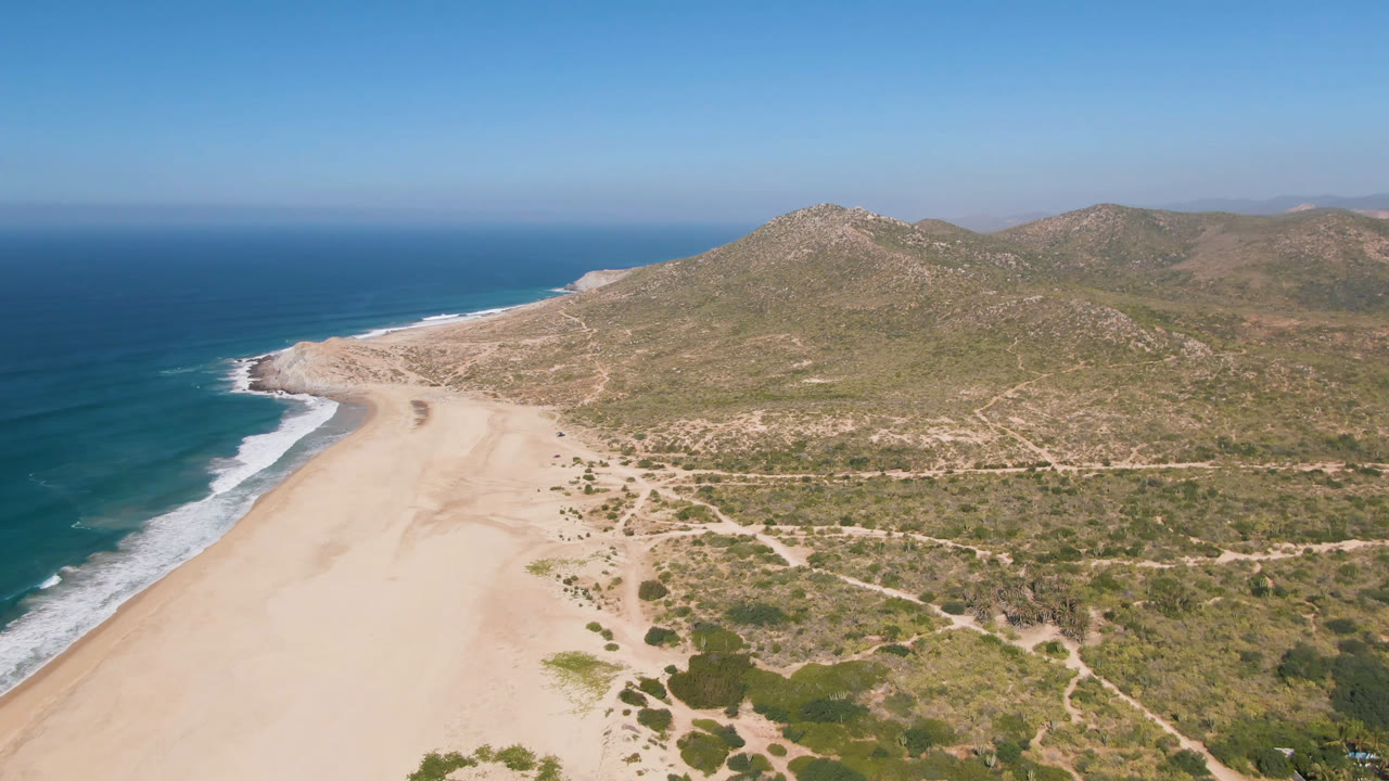 Opening aerial frame over Migrino, Baja California Sur, showing blue ocean surf beside a wide sandy beach, dry scrubland, dirt tracks, rocky hills, and no visible people.