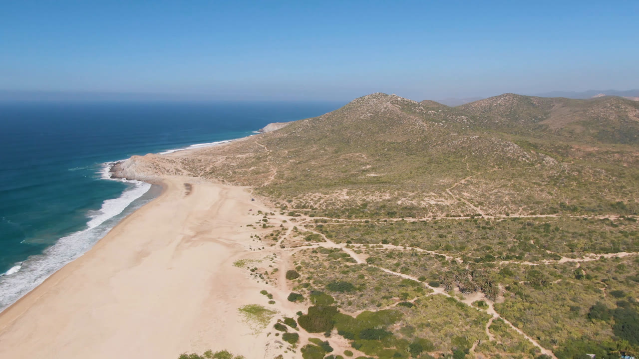 Aerial frame over Migrino beach with the curving shoreline on the left, pale sand through the middle, scrub and dirt routes in front, dry hills behind, and no visible people.