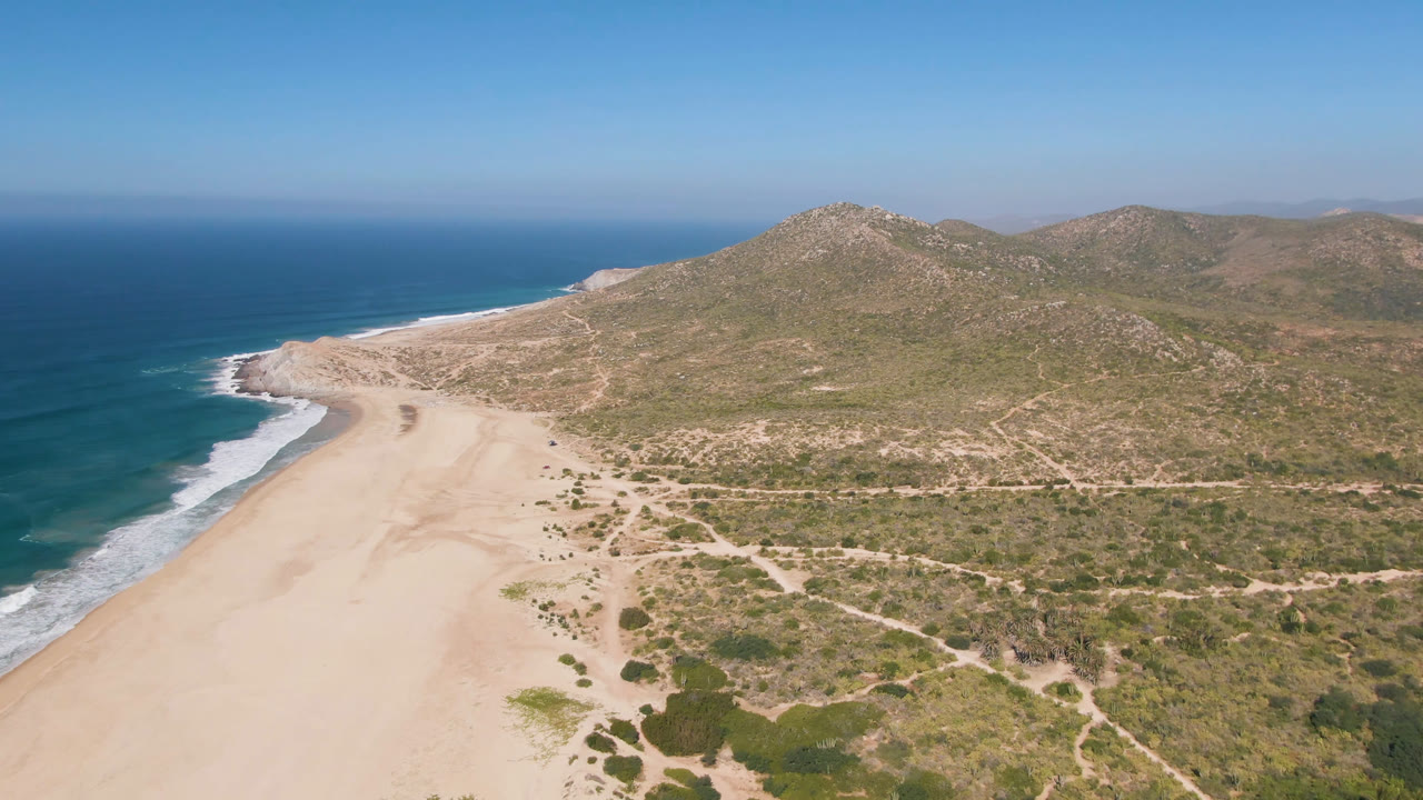 High coastal aerial with ocean, pale beach, scrubby flats, and mountains beneath clear sky.