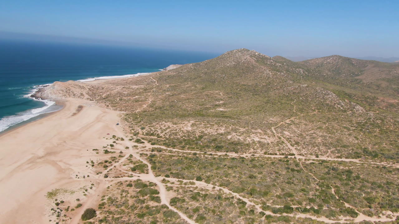 Wide aerial frame over Migrino, Baja California Sur, with pale beach sand at left, white surf along the ocean edge, scrub-covered ground, rocky hills beyond, and no visible people.