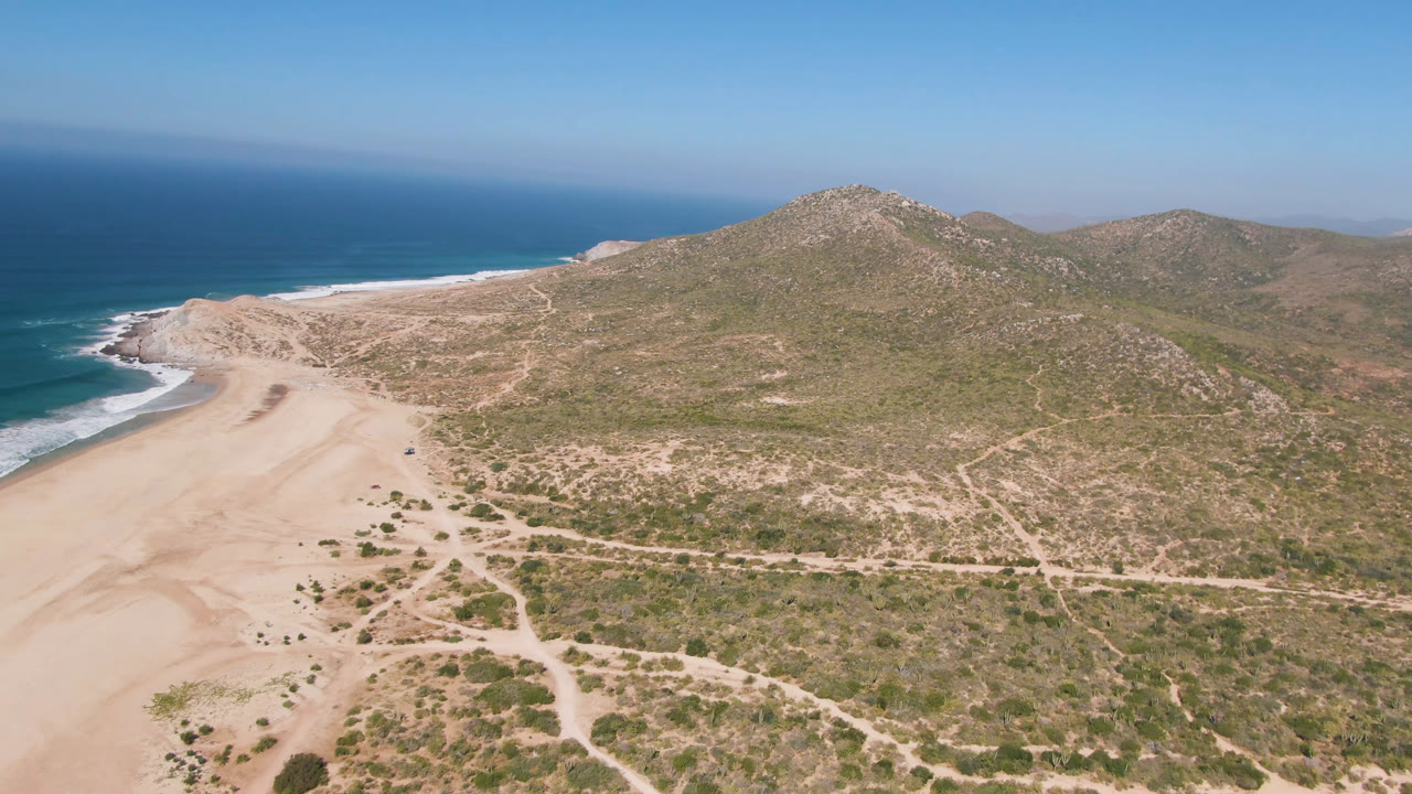 Aerial view over Migrino showing the surf line curving along the left, open sand in the lower left, desert scrub and dirt tracks across the center, rocky hills in back, and no visible people.
