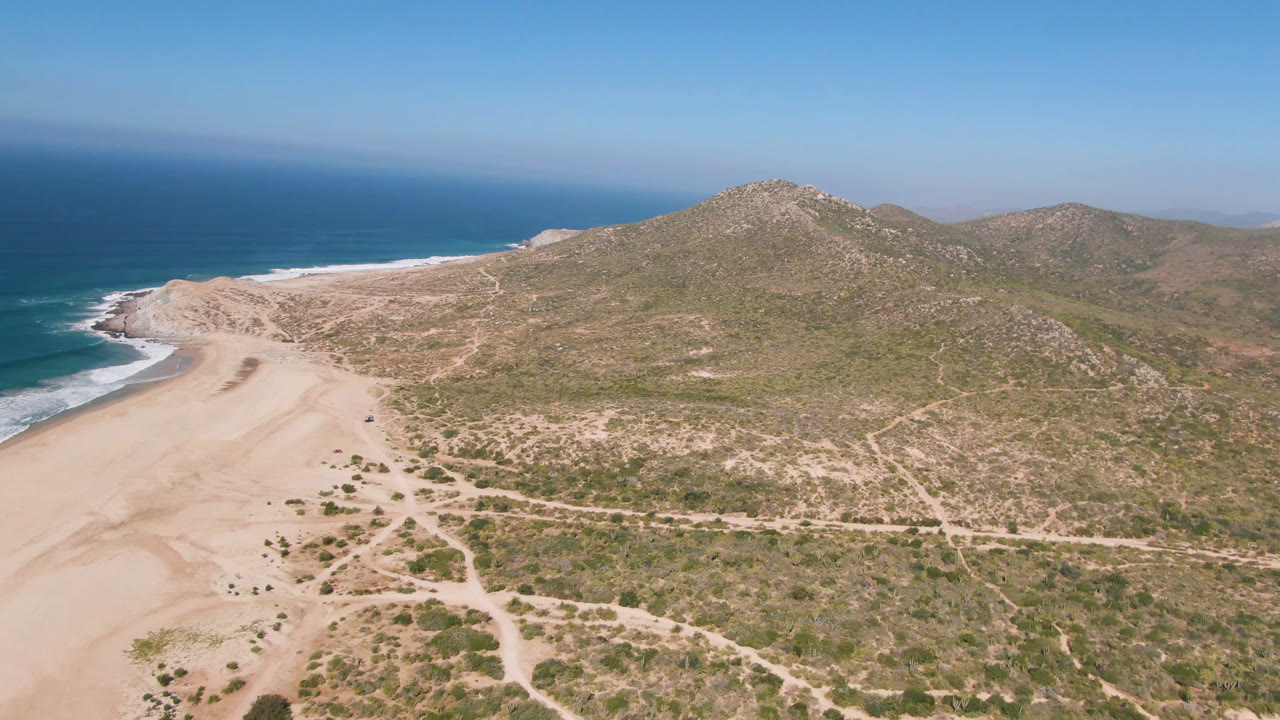 Drone view of beach dunes, scrubland, ocean, and low mountains under blue sky