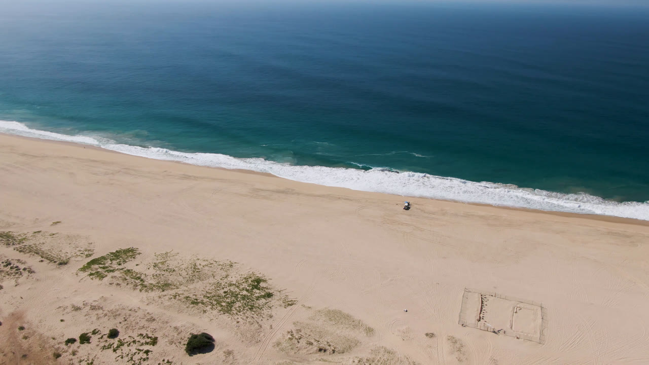 Aerial view over Migrino, Baja California Sur, with dark blue ocean, a white surf line, open sand, low dune vegetation, and no visible people.
