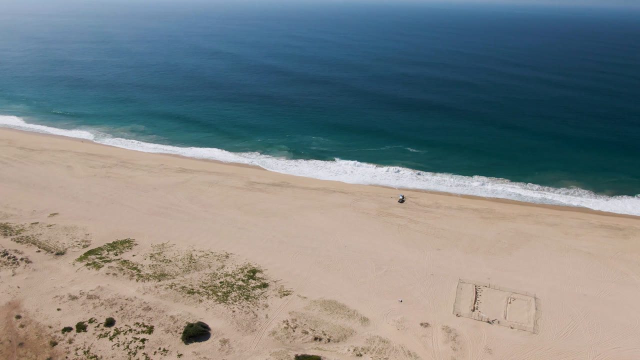 One second into the clip, the Migrino beach remains open and uncrowded, with the surf line across the upper third and pale sand filling the lower frame.