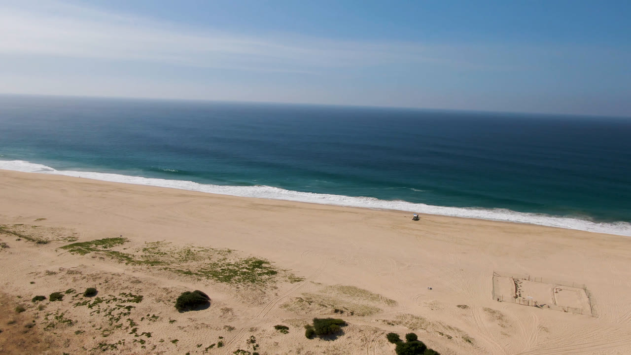 Opening aerial frame over Migrino, Baja California Sur, with a sand-marked soccer field in the lower right, scrubby dunes in front, white surf beyond the beach, and no recognizable people visible.
