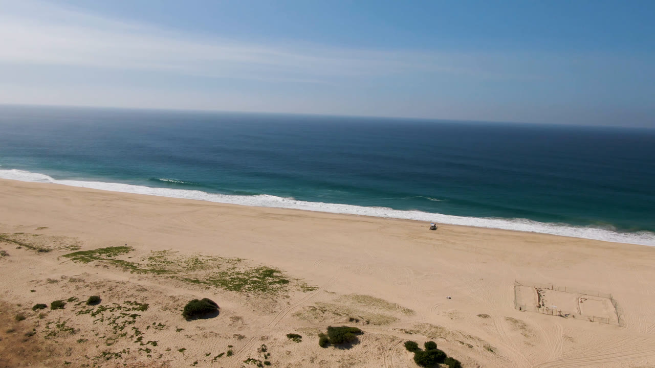 Aerial view in Migrino showing the rectangular soccer field etched into the sand near the lower right, scattered green shrubs across the dunes, a wide empty beach, and rolling surf.