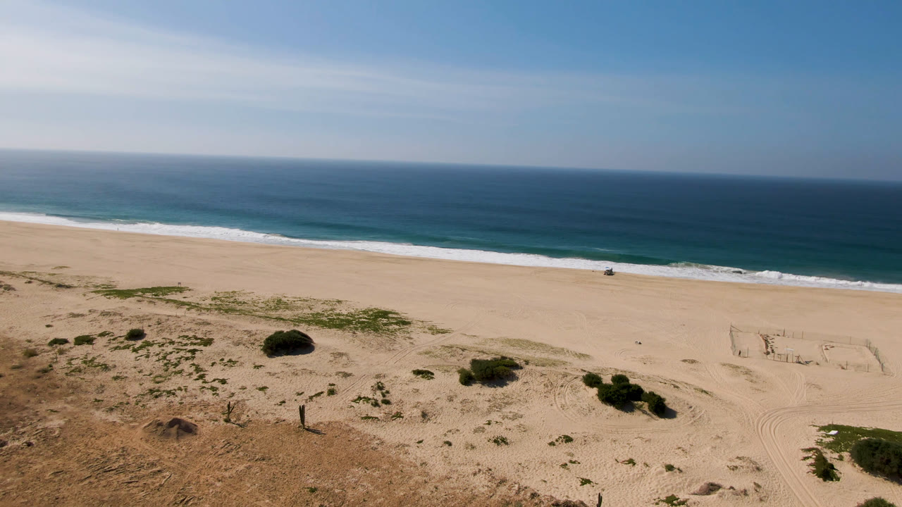 Migrino beach frame with the soccer field slightly right of center, tire tracks across the sand, low vegetation in the foreground, ocean surf behind, and no visible people.