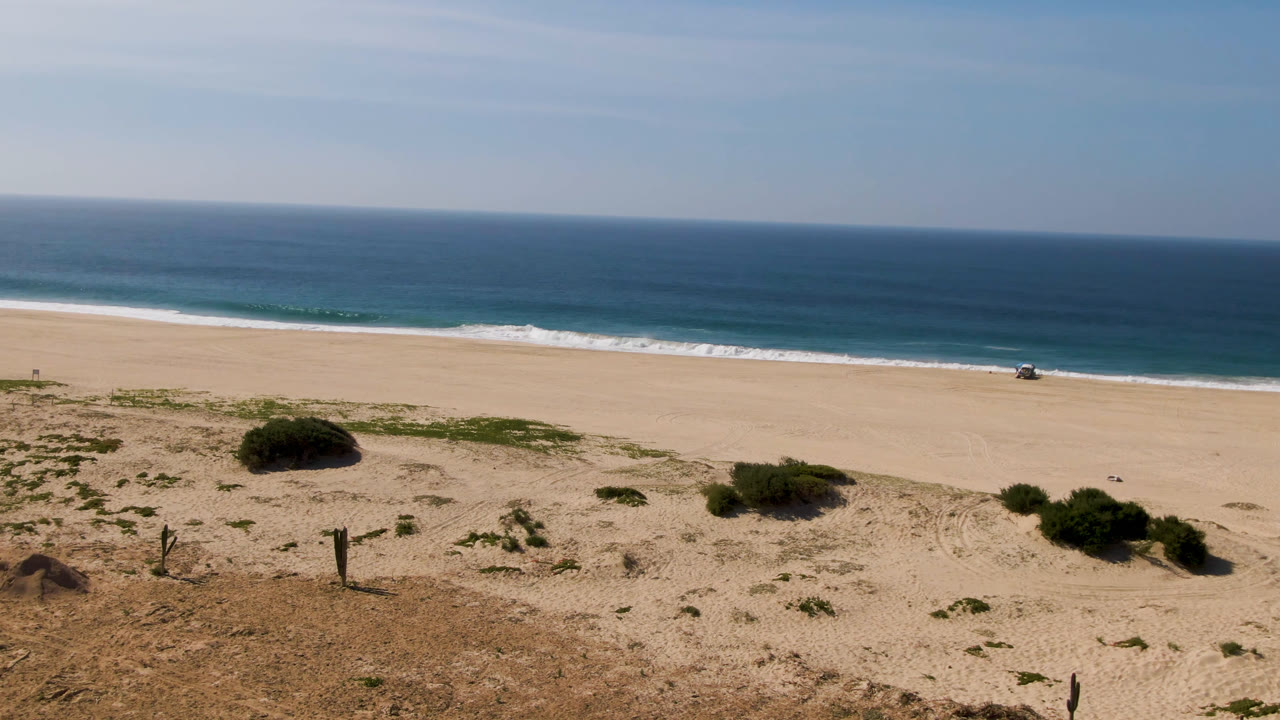 Wide Migrino shoreline frame with tan sand across the middle, scattered scrub in front, small surf lines in back, and no clearly identifiable people.